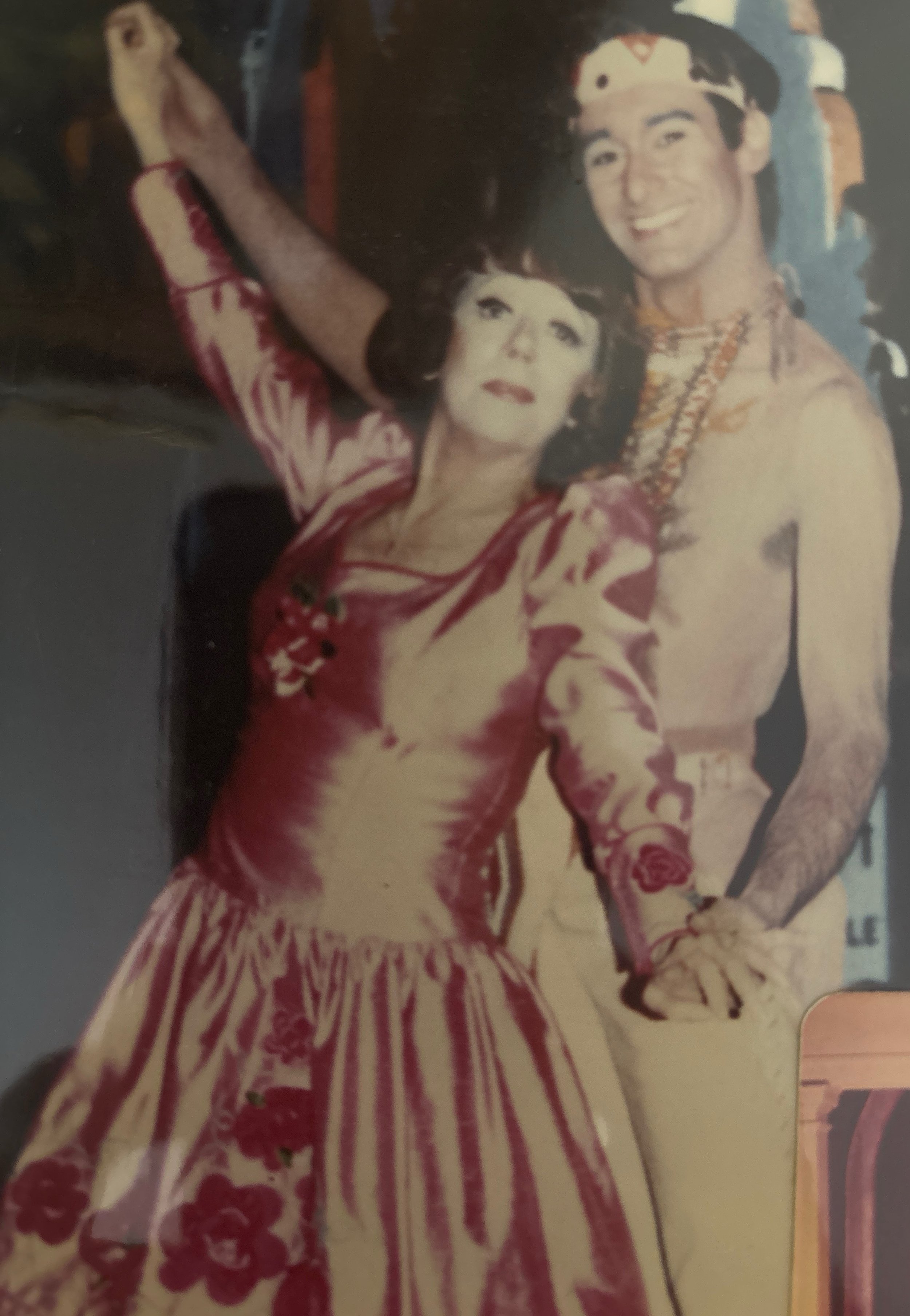 A young man wearing an animal print crown, an orange Thai traditional shirt, and a beaded necklace, smiling and posing next to a woman in a vintage-style dress with a floral pattern, in an indoor setting.