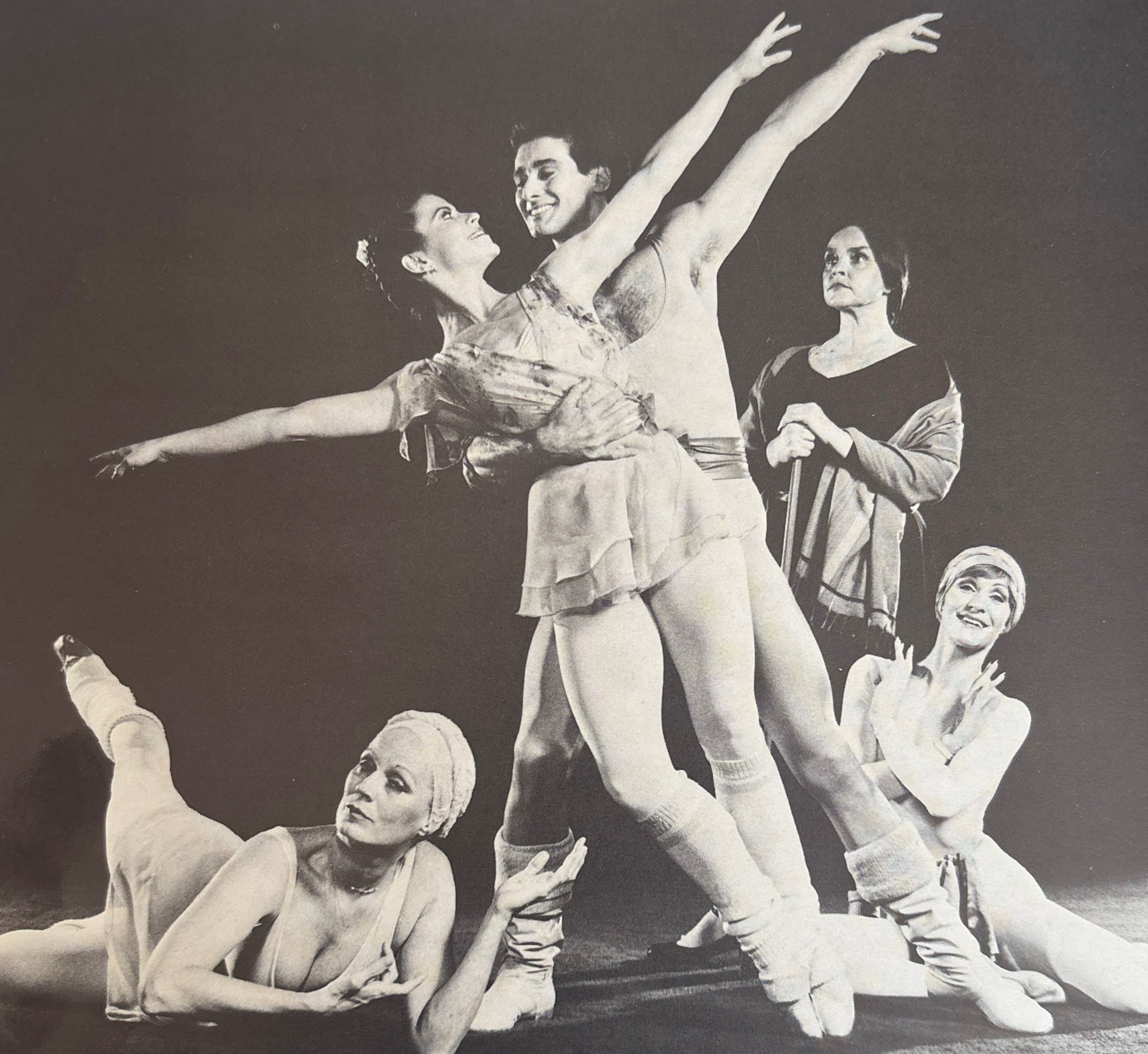 Black and white photo of five women dancing, with their arms raised, wearing matching outfits, performing a choreographed dance routine.