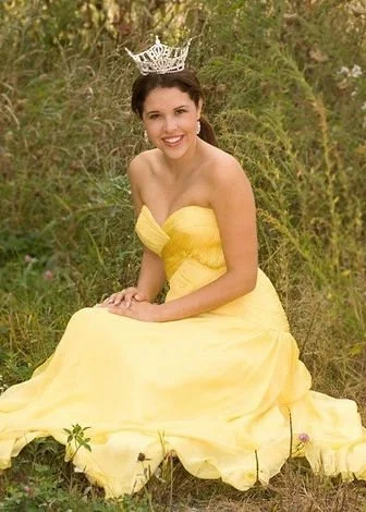 A young woman sitting outdoors on grass, wearing a yellow strapless dress and a tiara, smiling at the camera.