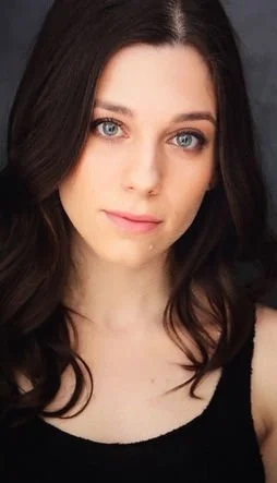 Close-up of a young woman with fair skin, long dark hair, blue eyes, and a neutral expression, wearing a black top.