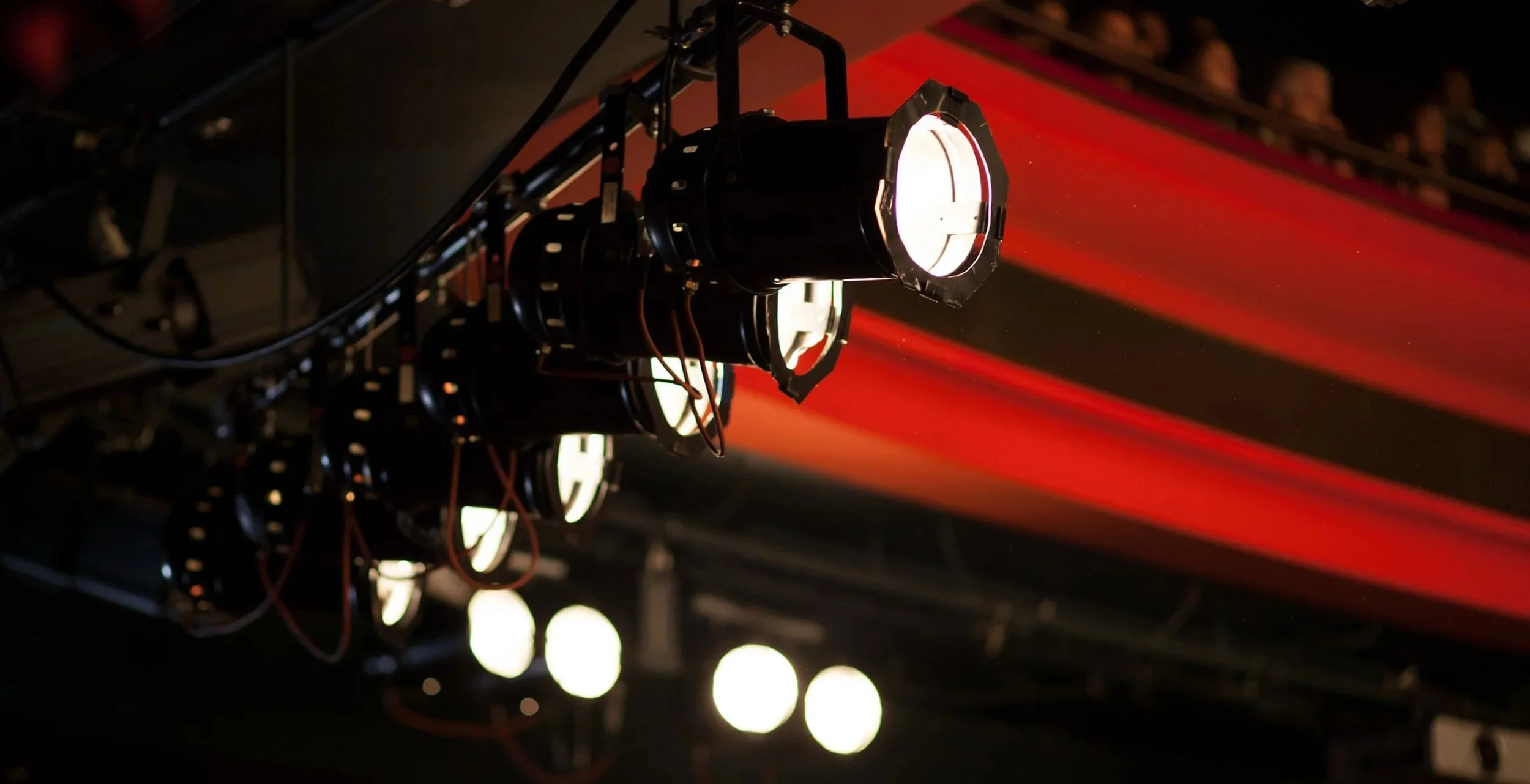 Stage lighting fixtures with bright white and red lights, mounted on a metal rigging, with red light beams projecting in the background.