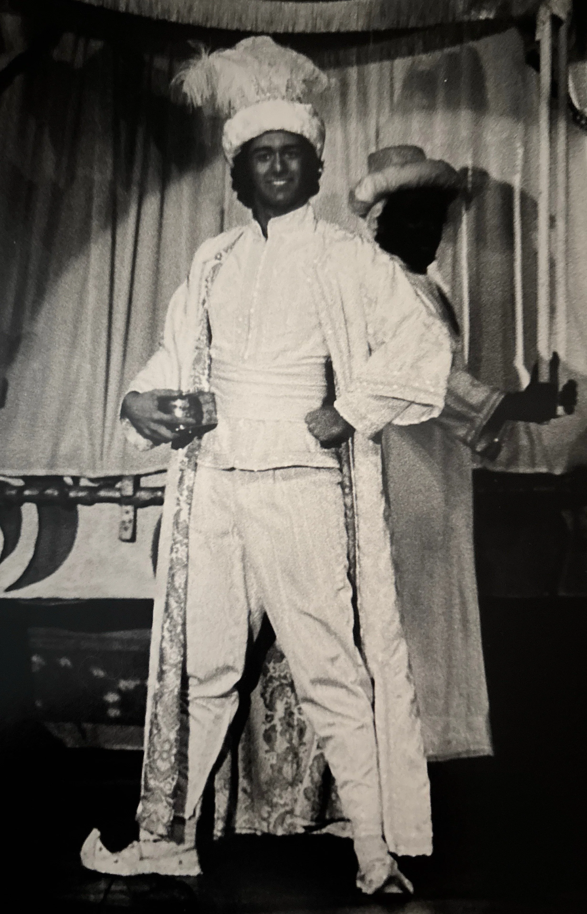 A person dressed in traditional Filipino attire, wearing a white barong tagalog and a large feathered headdress, standing on stage with a curtain in the background.