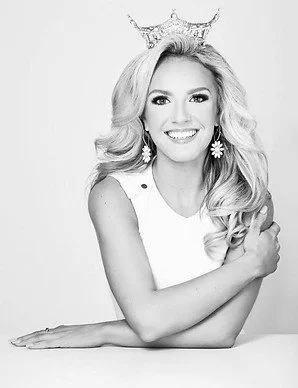 Black and white photo of a woman with long wavy hair, smiling and wearing a crown and earrings.