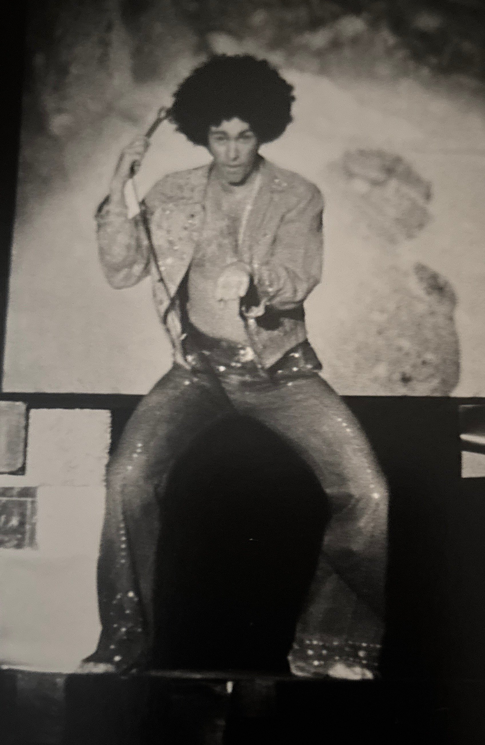 A person with a large afro hairstyle and a patterned jacket holding a hairbrush, sitting on the ground in front of a black and white poster of themselves.