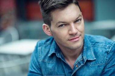A young man with short dark hair wearing a blue denim shirt, looking slightly to the side with a subtle smile, outdoors in an urban setting.
