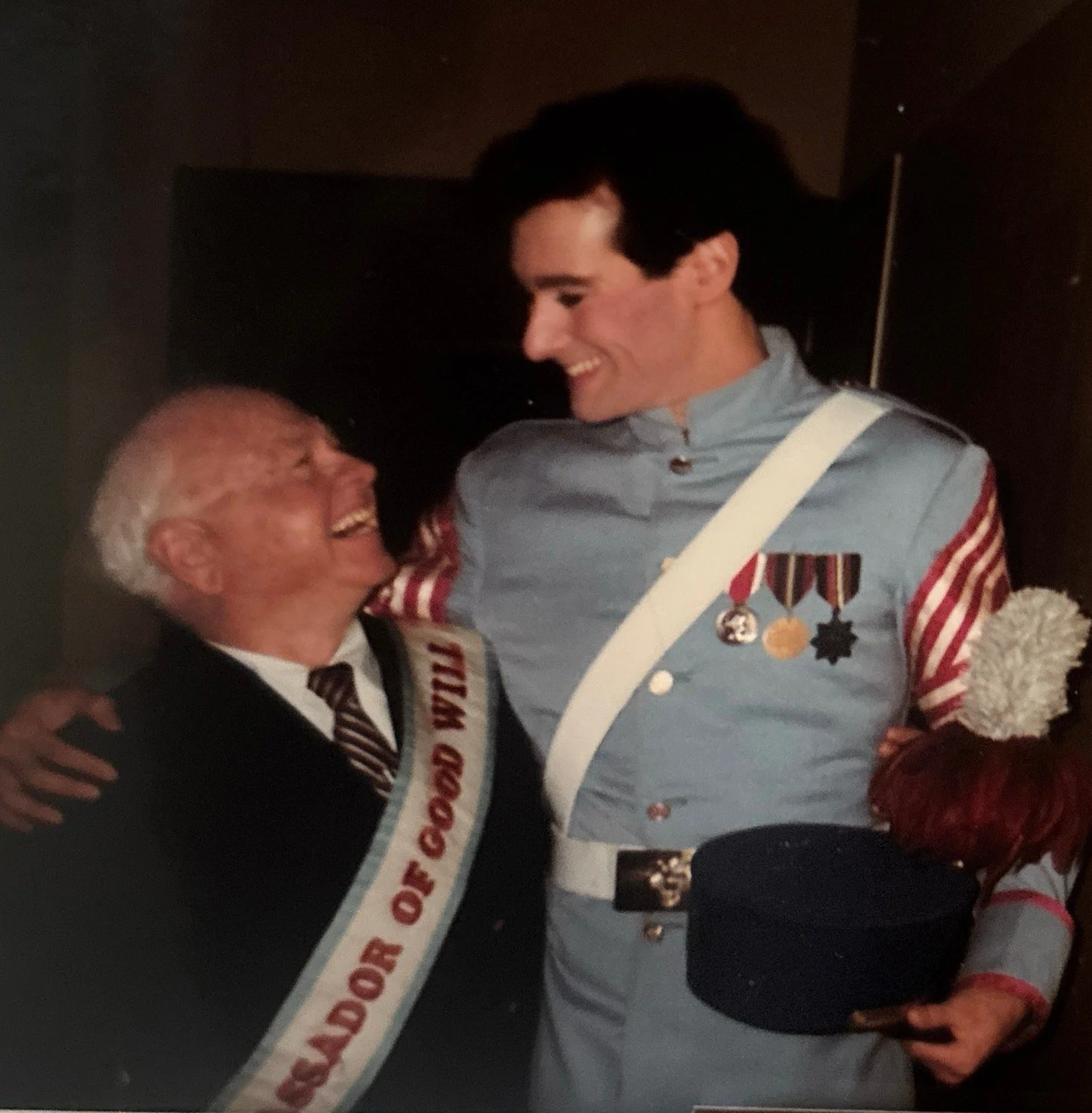 A young man in a band uniform with medals on his chest, smiling as he embraces an older man dressed in a dark suit and tie, both smiling warmly.