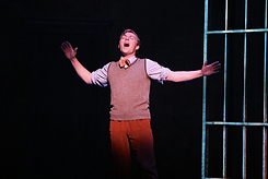 A person singing on stage with arms outstretched, wearing a beige sweater vest over a white shirt with rolled sleeves, and red pants, standing next to a metal barred structure.