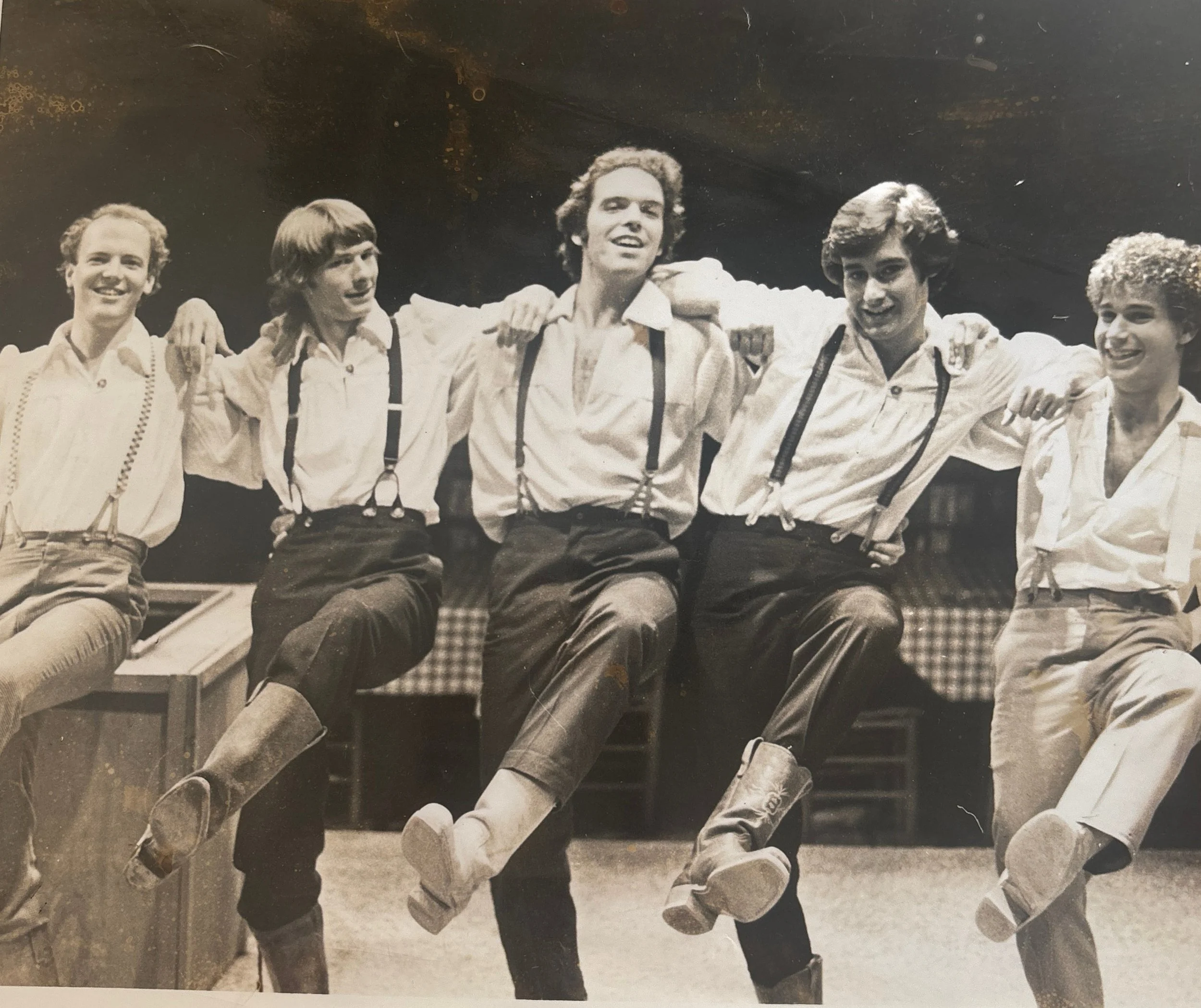 A black-and-white photo of five young men sitting together with their arms around each other's shoulders, smiling and looking at the camera, dressed in casual clothing with suspenders and white shirts.