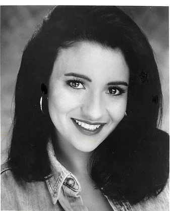 Black and white portrait of a young woman with shoulder-length dark hair, smiling, wearing hoop earrings and a denim jacket.