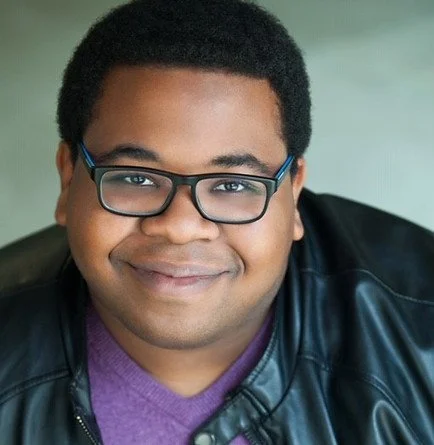 Close-up of a young man with glasses smiling, wearing a purple shirt and black leather jacket.