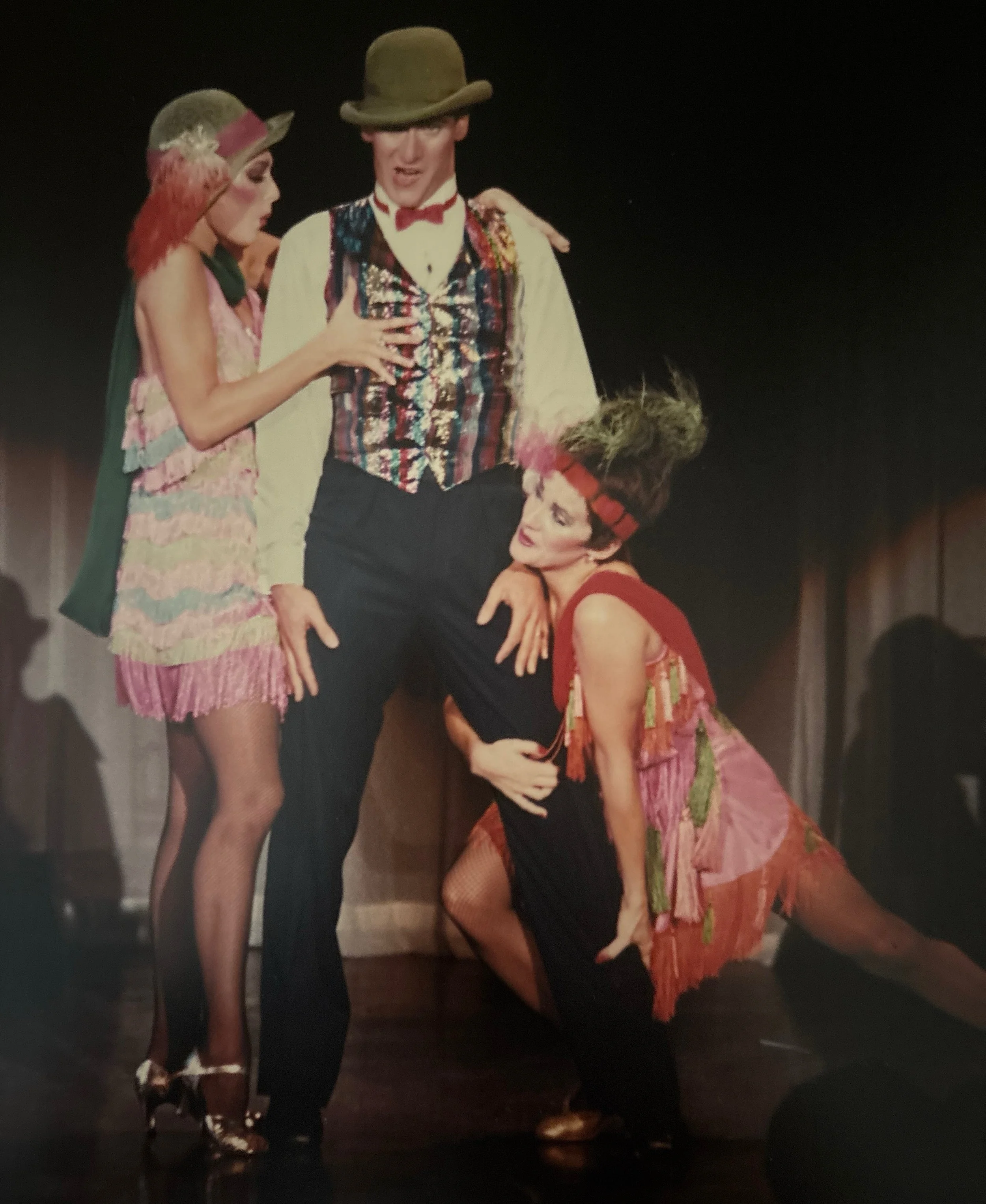 Four performers on stage dressed in colorful costumes, two women in pink and green gowns with hats, and two men in vests and hats, one standing and one kneeling, engaged in a theatrical scene.