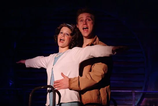 Two actors performing on stage, with the woman in a white cardigan and the man in a tan jacket, both looking joyful.
