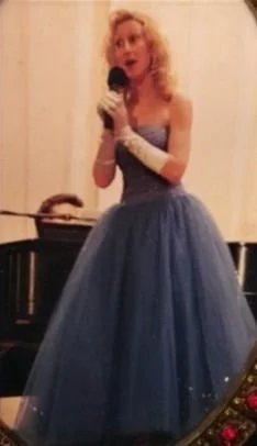 A woman in a strapless blue ball gown holding a microphone, standing in front of a piano.