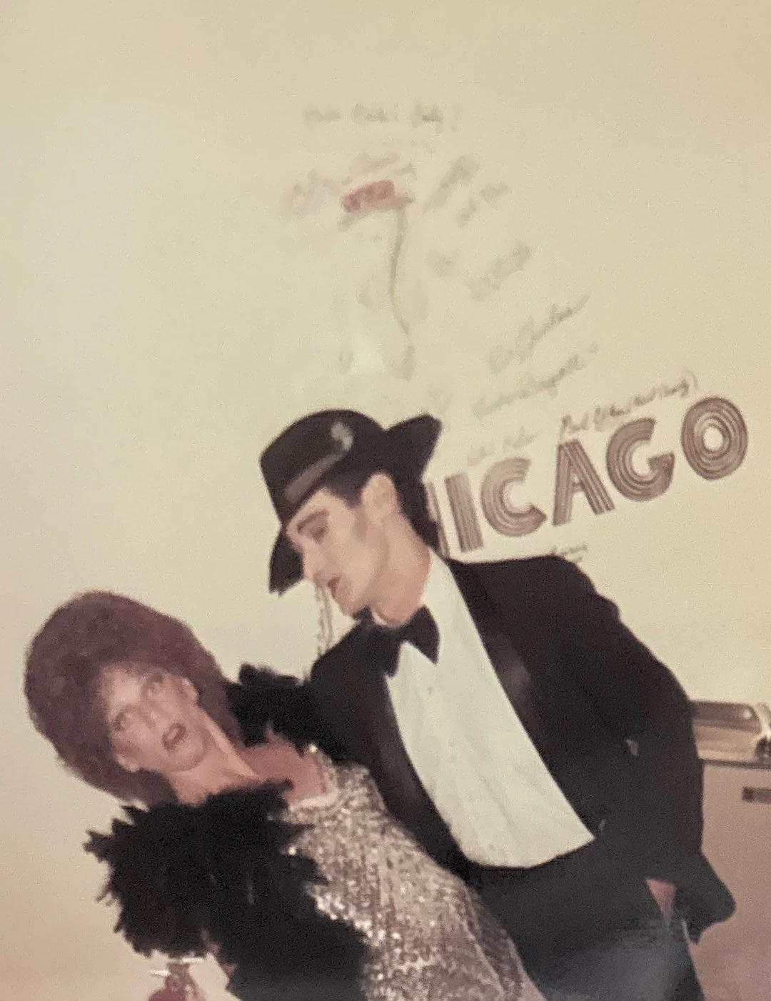 A man dressed as James Bond, wearing a tuxedo and fedora, is smiling and dancing with a woman in a sparkly dress in front of a white wall with vintage Chicago-themed artwork.