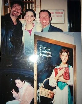 Group of three people posing together, with two framed photos of a woman holding a book or a basket in the background.
