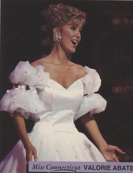 A woman in a white, Victorian-style dress with puffy sleeves and a full skirt, smiling on stage with a dark background.
