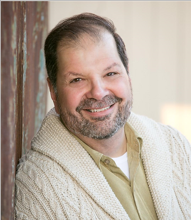 A smiling man with dark hair and beard posing indoors, wearing a beige knit sweater and a collared shirt.