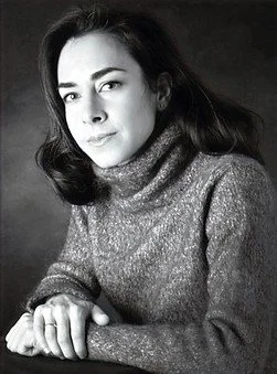 Black and white portrait of a woman with dark hair, wearing a turtleneck sweater, looking at the camera with a neutral expression, with her hands folded on a surface.