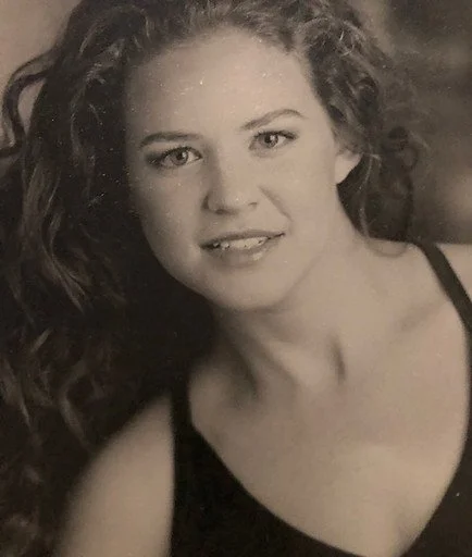 A black and white portrait of a young woman with curly hair, wearing a tank top, smiling softly at the camera.