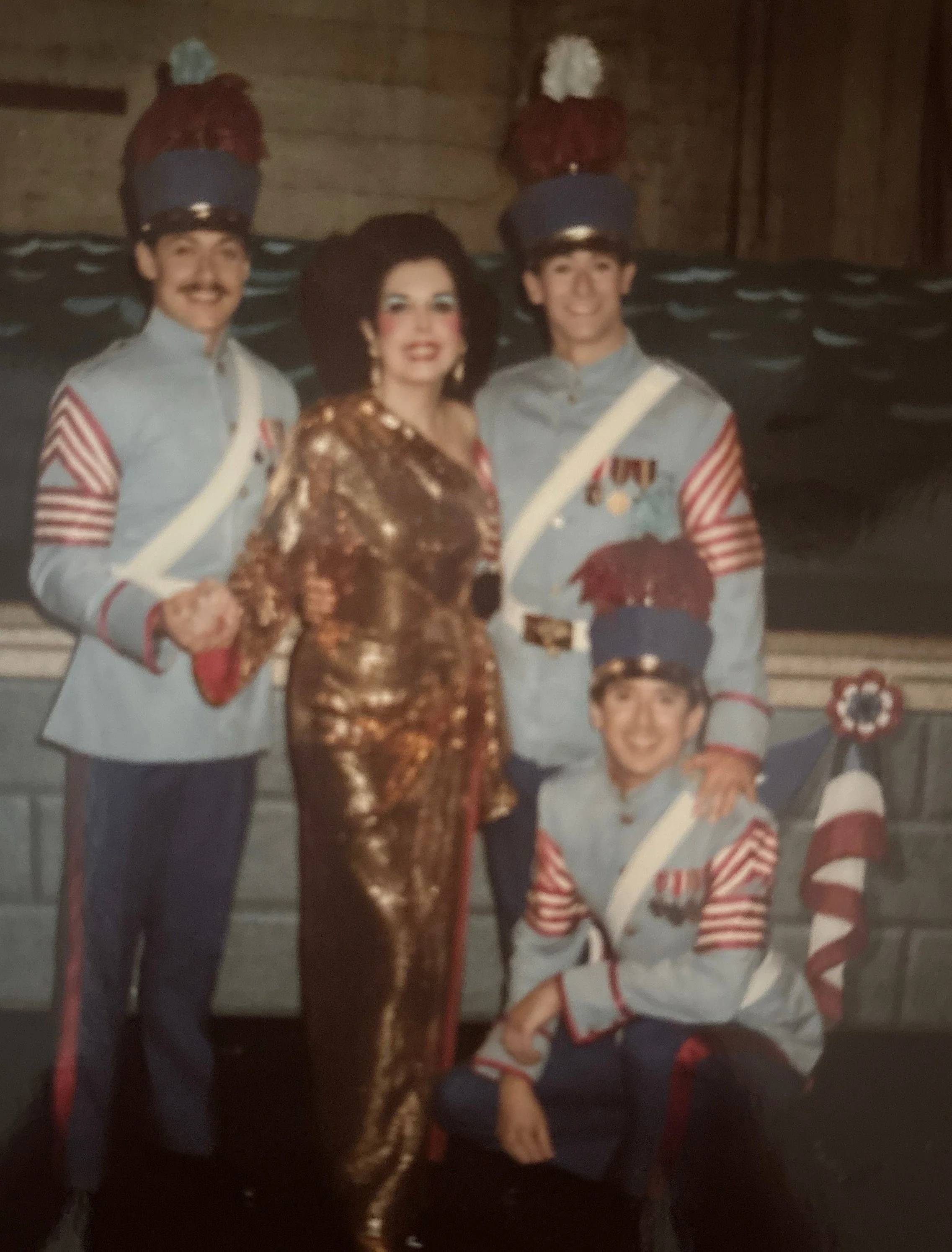 Two men dressed in military uniforms with blue hats and red plumes, a woman in a shiny gold dress and large black hat, and a boy in a scout uniform. They are posing together inside a building with wood paneling in the background.