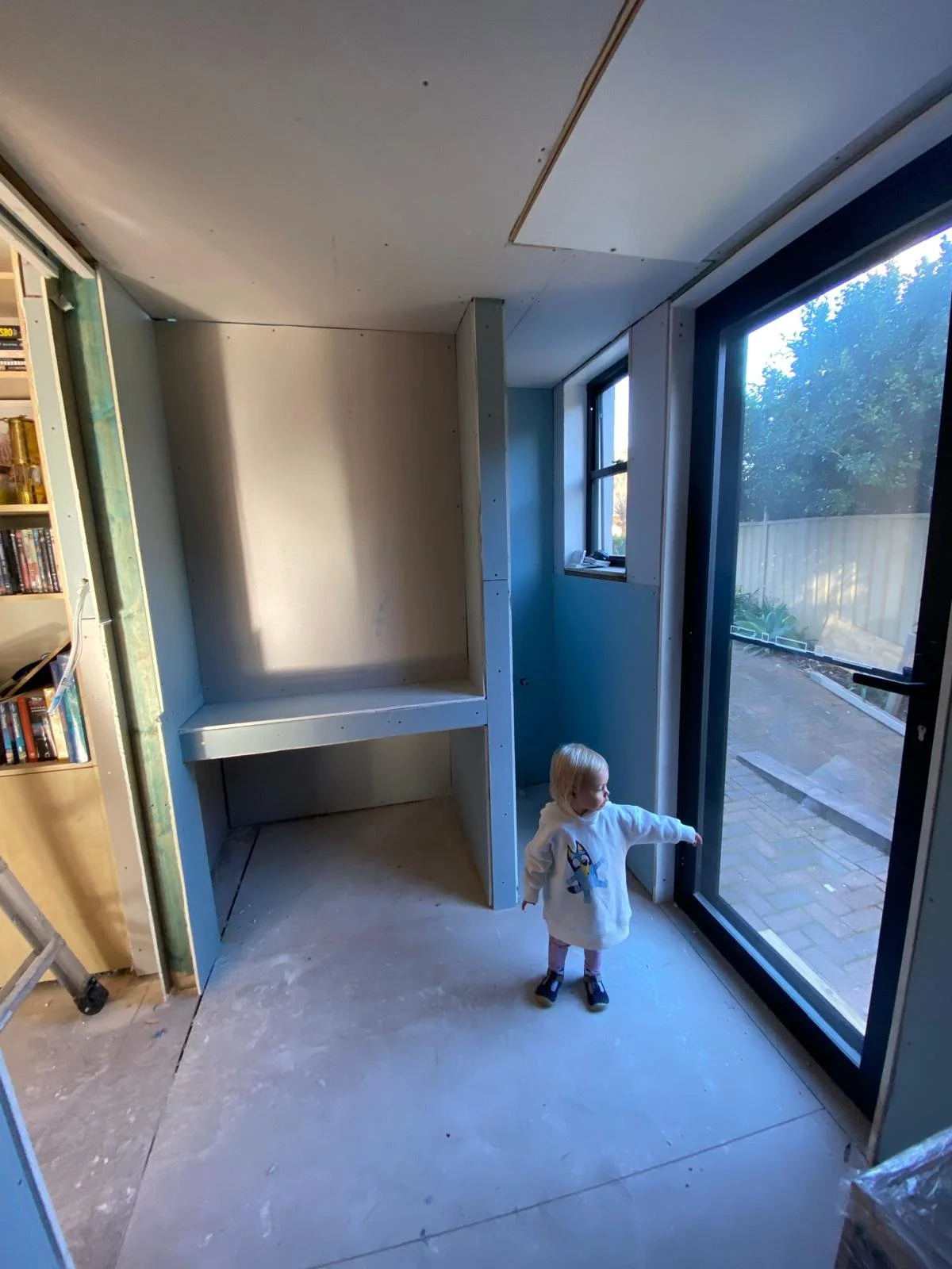 A small child with blonde hair wearing a white hoodie with a cartoon character on it, standing inside a house under construction or renovation, pointing towards the large sliding glass door leading outside. The interior is partially finished with visible drywall and a bookshelf on the left.