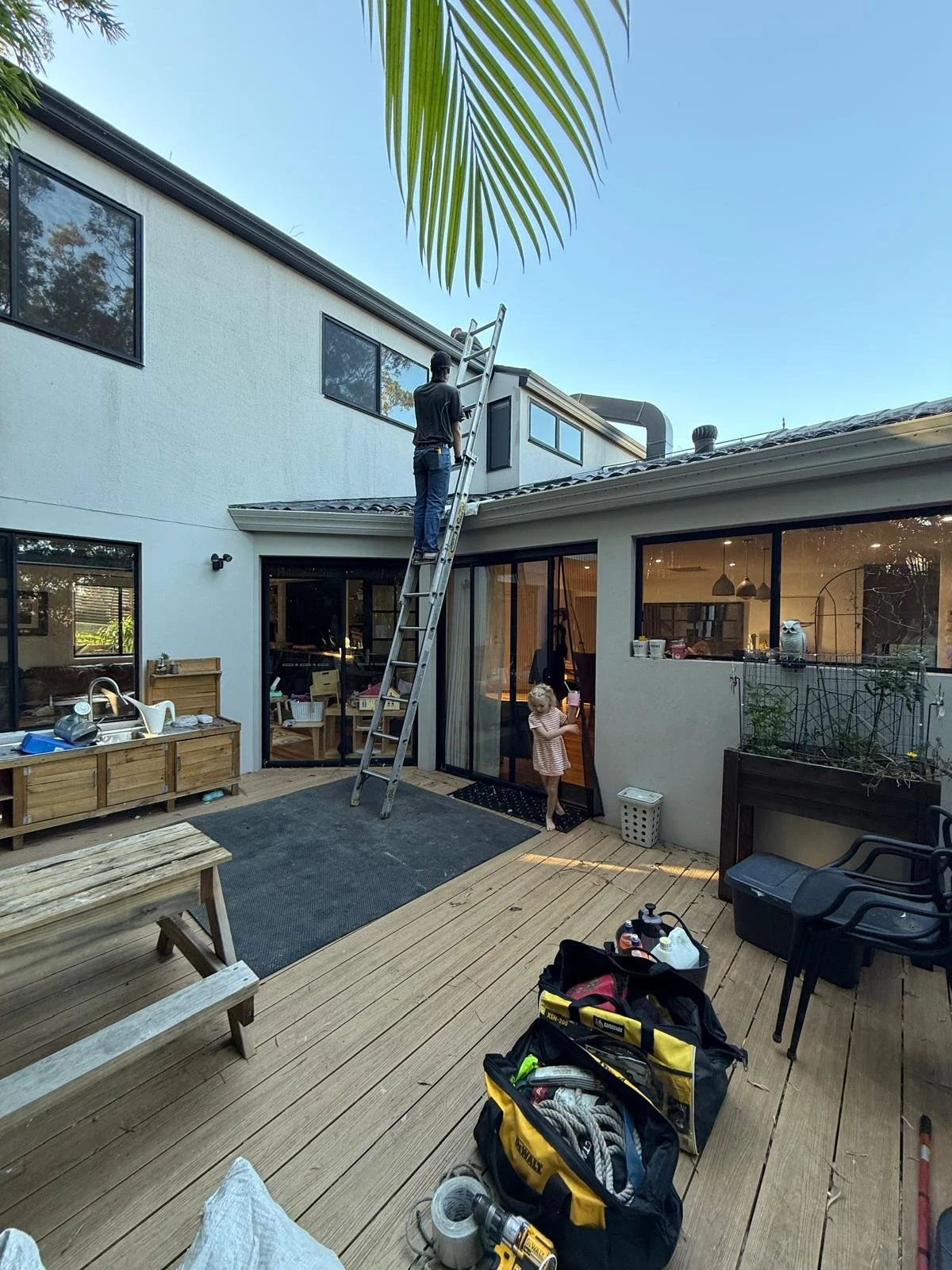 A man on a ladder working on the exterior of a modern two-story house with large windows; a young girl stands nearby on a wooden deck, observing him; various tools and equipment are scattered around on the deck.