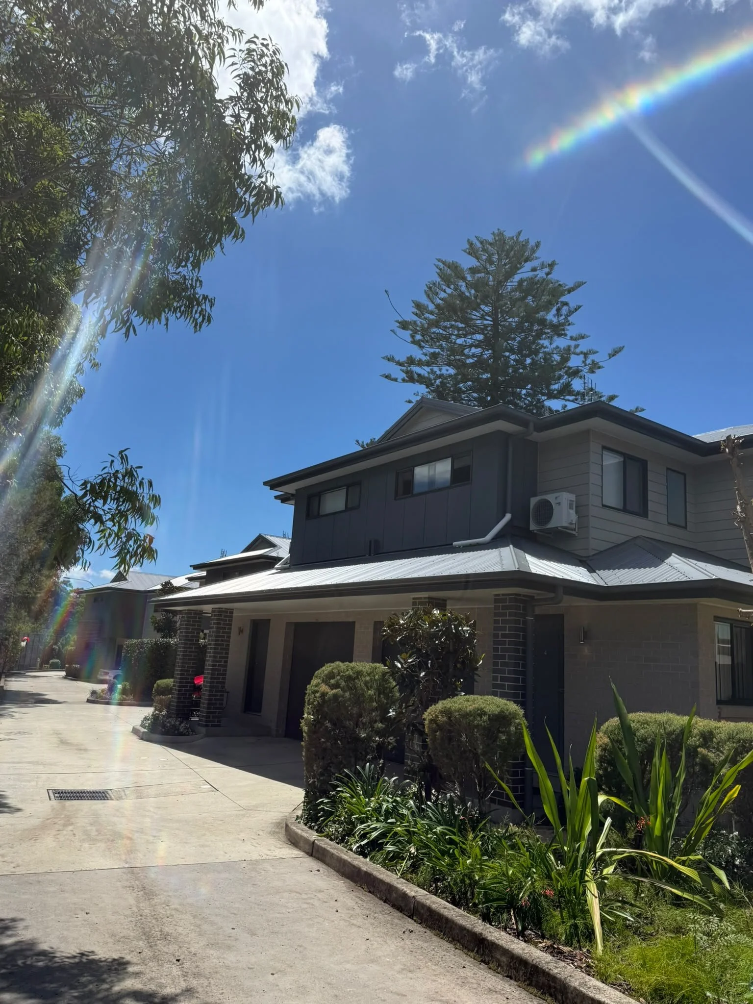 A modern residential building with a metal roof and brick walls; landscaping with bushes and plants; a driveway; a tall tree in the background; a blue sky with scattered clouds and a rainbow.