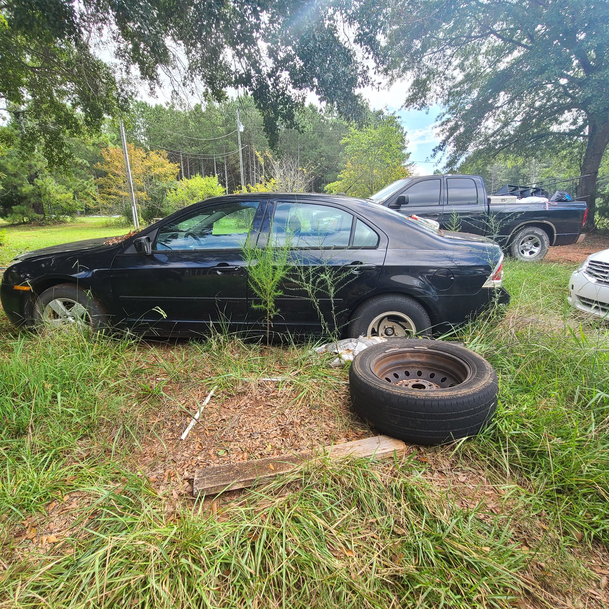Black sedan and black pickup truck parked on a grassy area surrounded by trees and utility poles, with a tire and wooden board on the ground nearby.