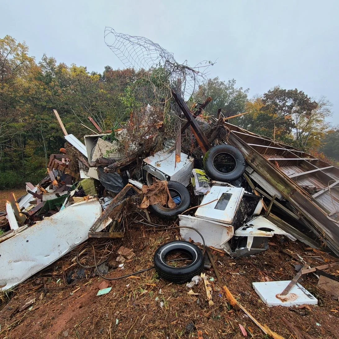 Debris and wreckage from a collapsed building, including broken appliances, tires, and metal sheeting, on a dirt ground with trees in the background.