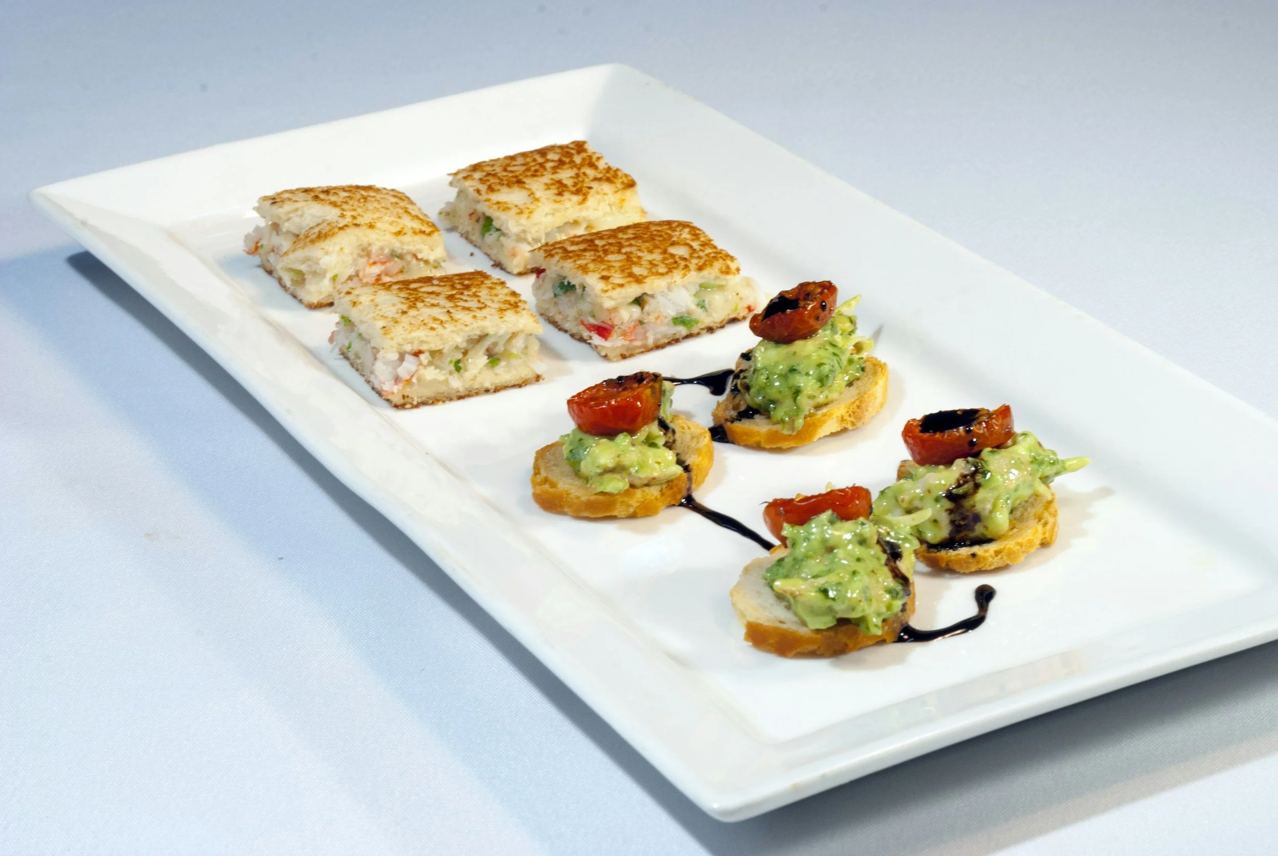 Appetizer platter with small sandwiches topped with avocado and cherry tomato, and square mini sandwiches with seafood and vegetables, garnished with balsamic glaze, on a white rectangular plate.