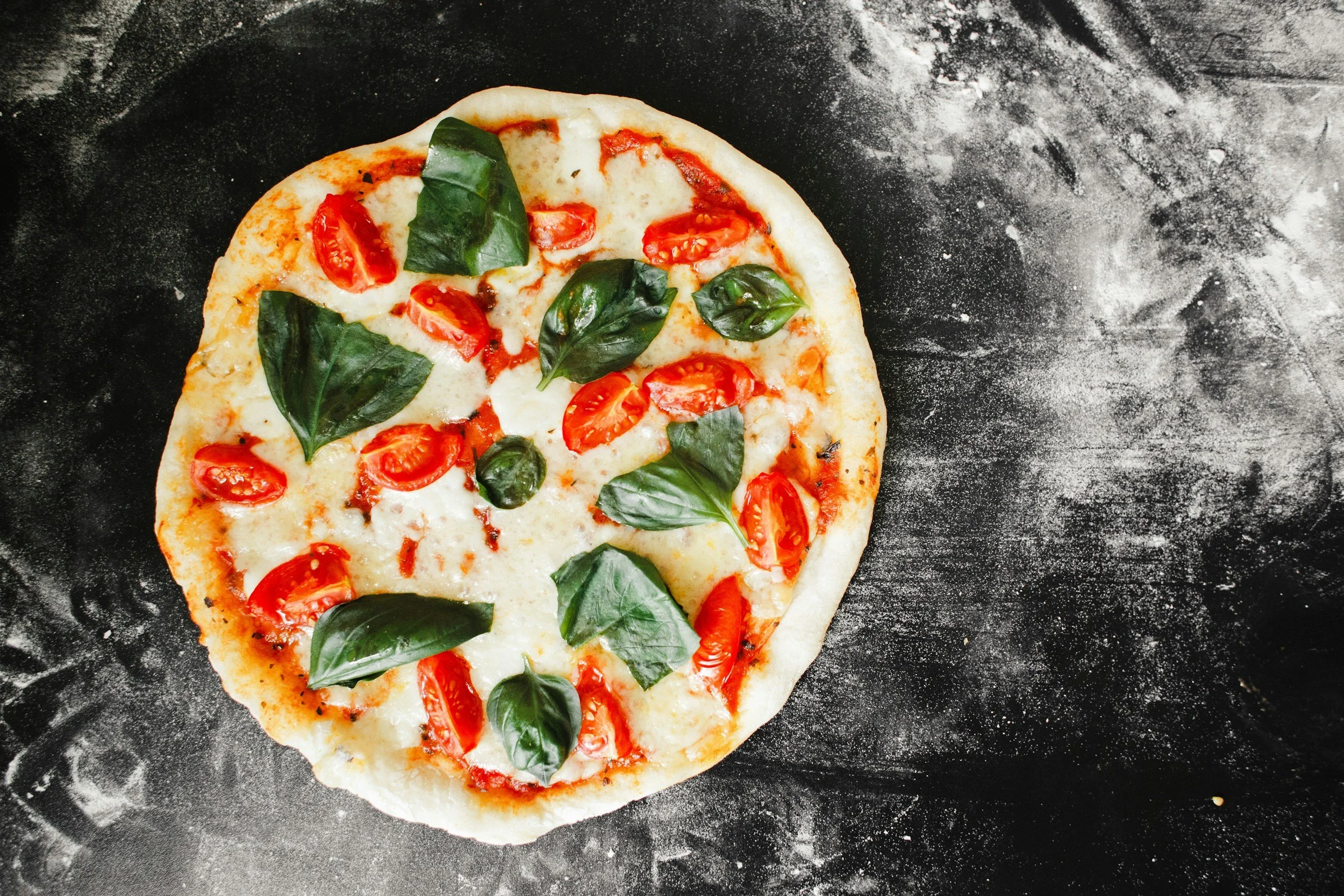 A close-up of a freshly baked pizza with cherry tomatoes and fresh basil leaves on a dark, floured surface.