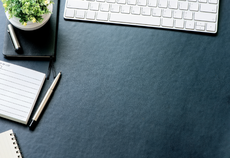 Top view of a desk with a white keyboard, a black notebook with a silver pen on top, an open notepad, a spiral notepad, and a small potted plant in a white pot.