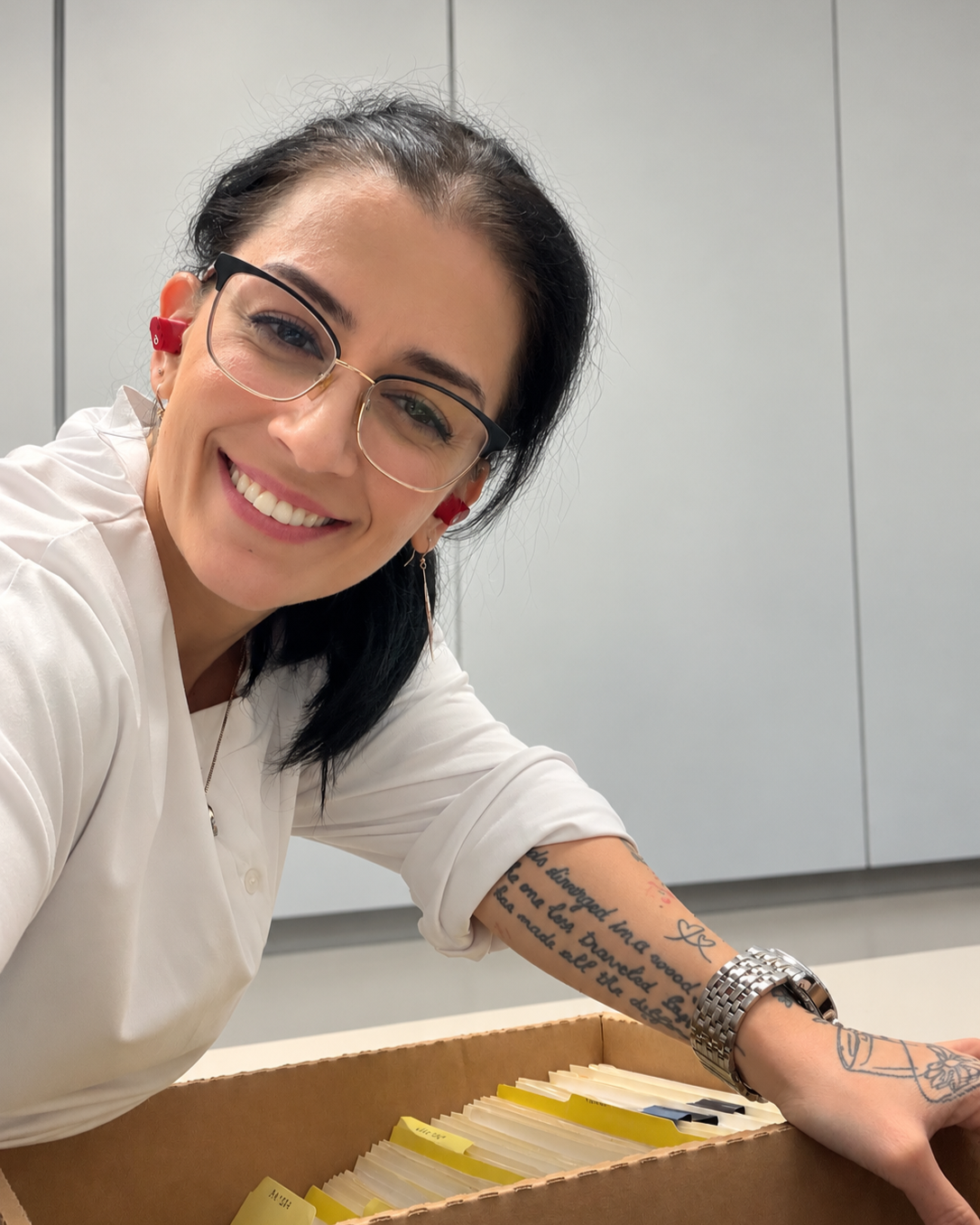 Mel Stepanian with glasses and tattoos on her arm, wearing a white shirt, leaning over a box of files with yellow labels, in a room with gray cabinets.