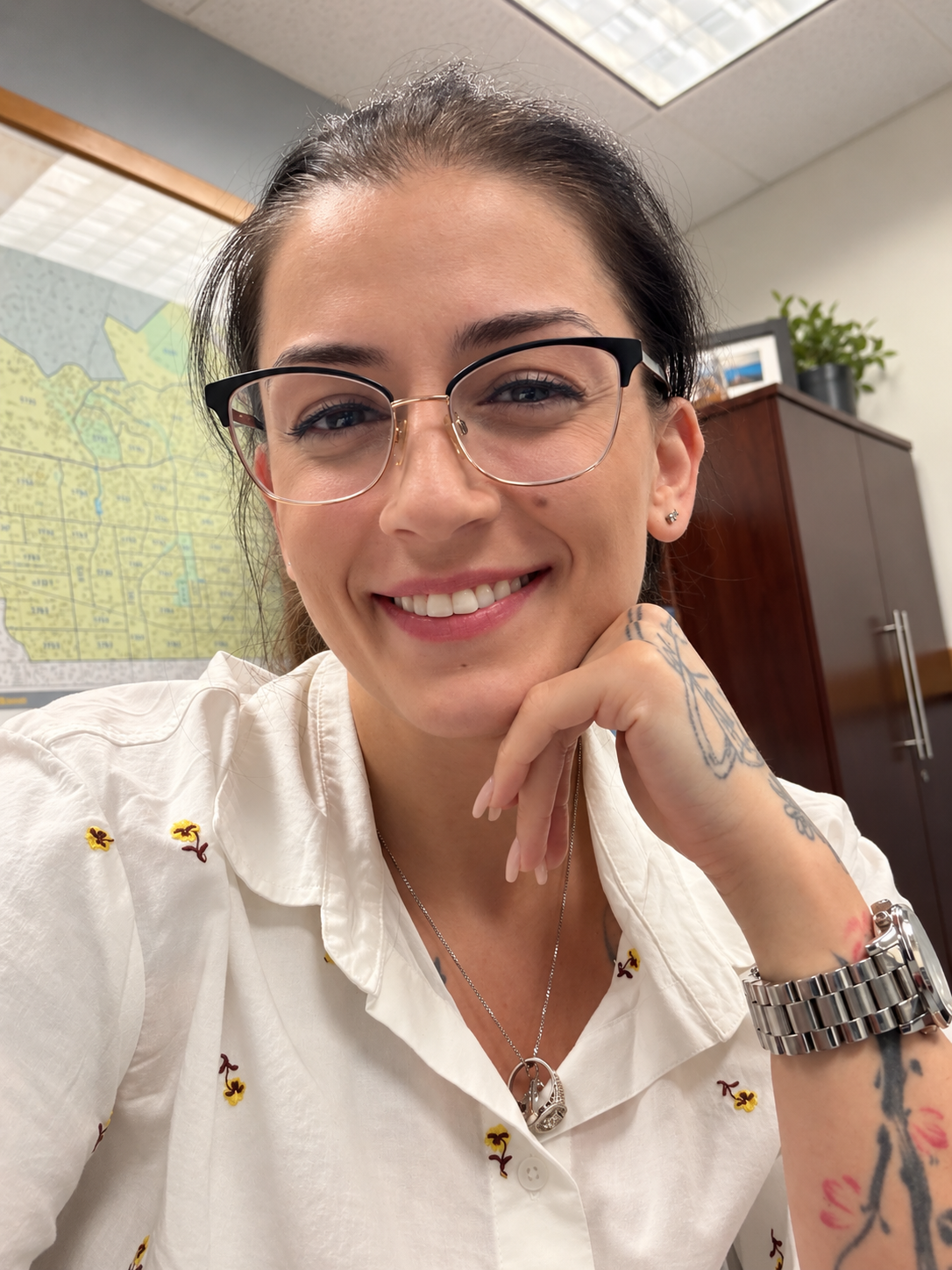 Close-up of a woman in an office setting smiling, wearing glasses, a white button-up shirt with small embroidered flowers, a silver watch, and a necklace with a pendant. She has tattoos on her hand and forearm, and a map is visible on the wall behind her.