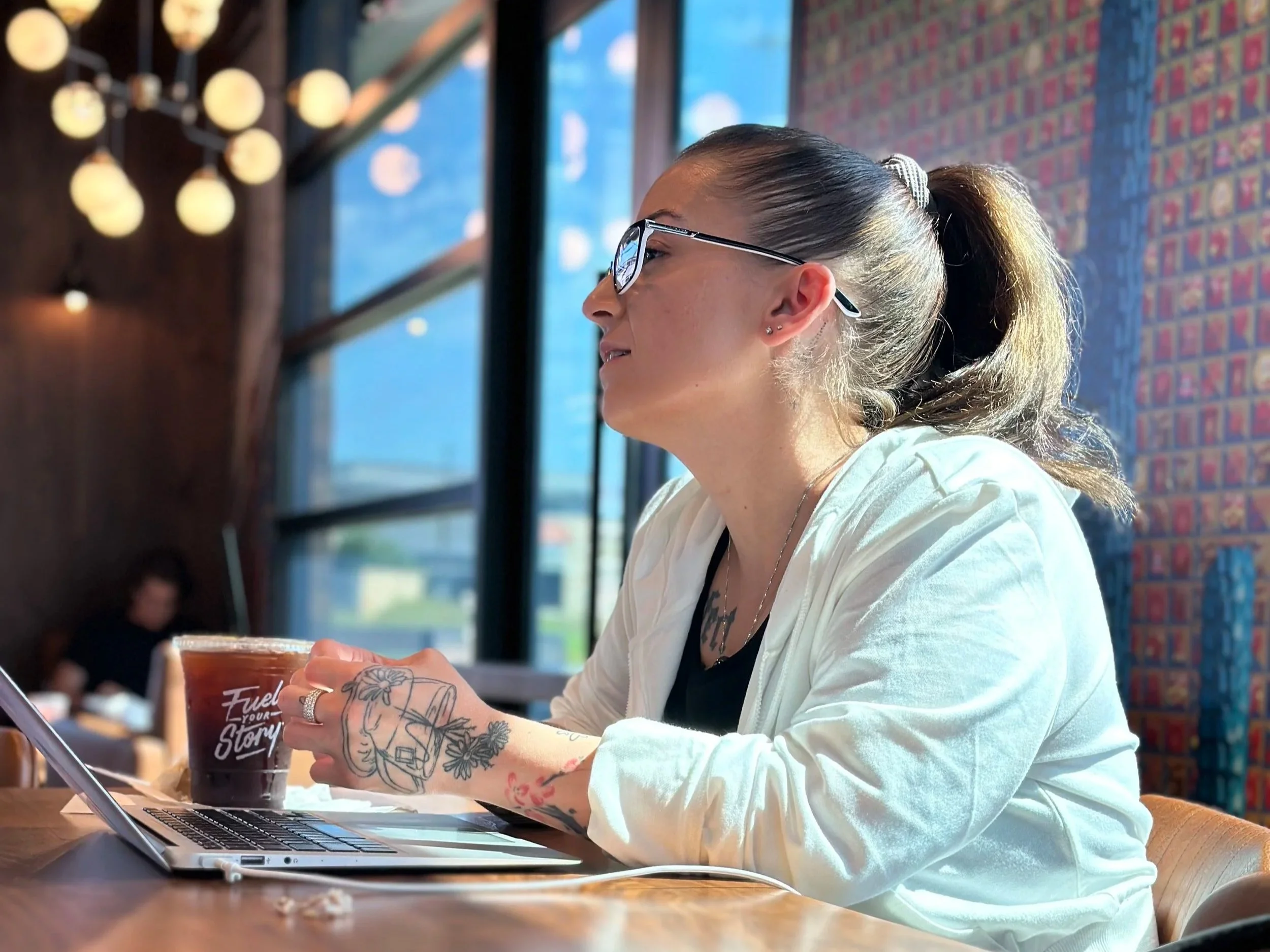 Mel Stepanian seated at a table in a coffee shop, holding a drink, with large windows and colorful brick walls behind her.