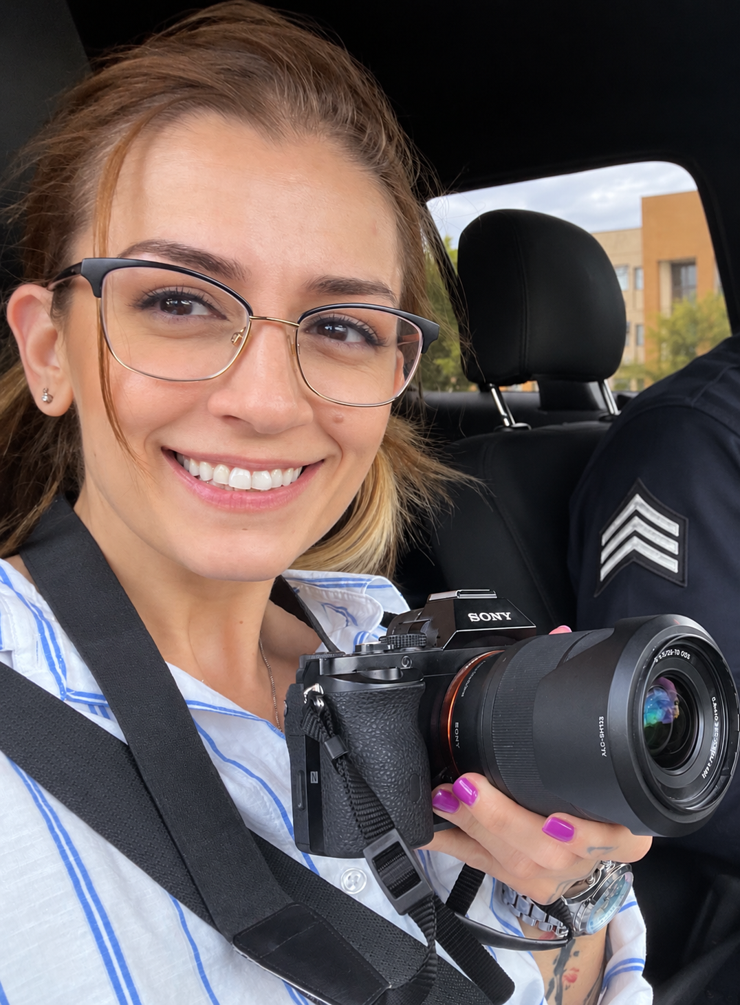 Mel Stepanian with glasses smiling, holding a Sony camera, seated in a vehicle with a police officer beside her.