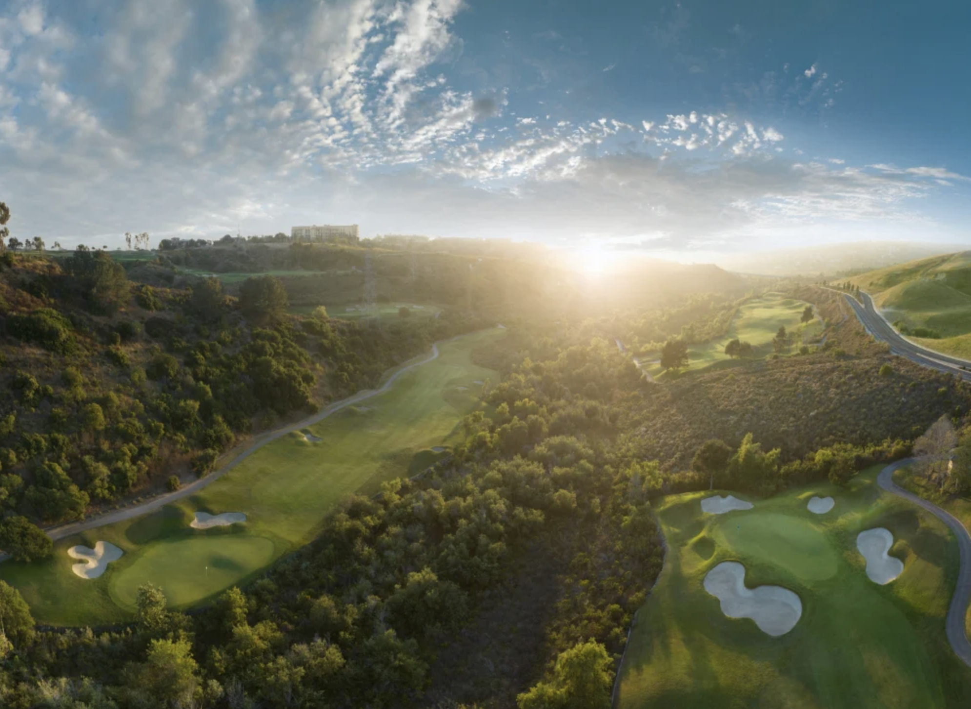Aerial view of a golf course surrounded by green trees and hills, with a road and a building visible in the background under a partly cloudy sky during sunset.
