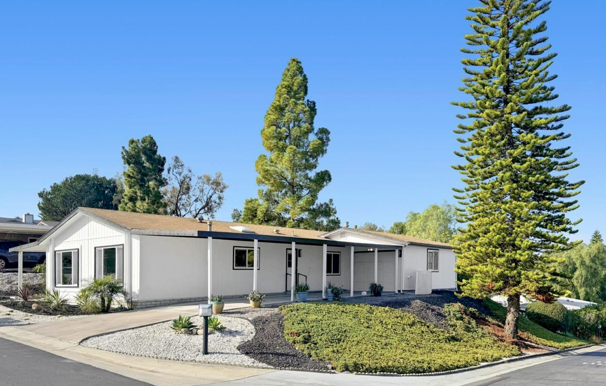 A white single-story house with a flat roof, surrounded by a landscaped yard with plants, trees, and gravel, under a blue sky.