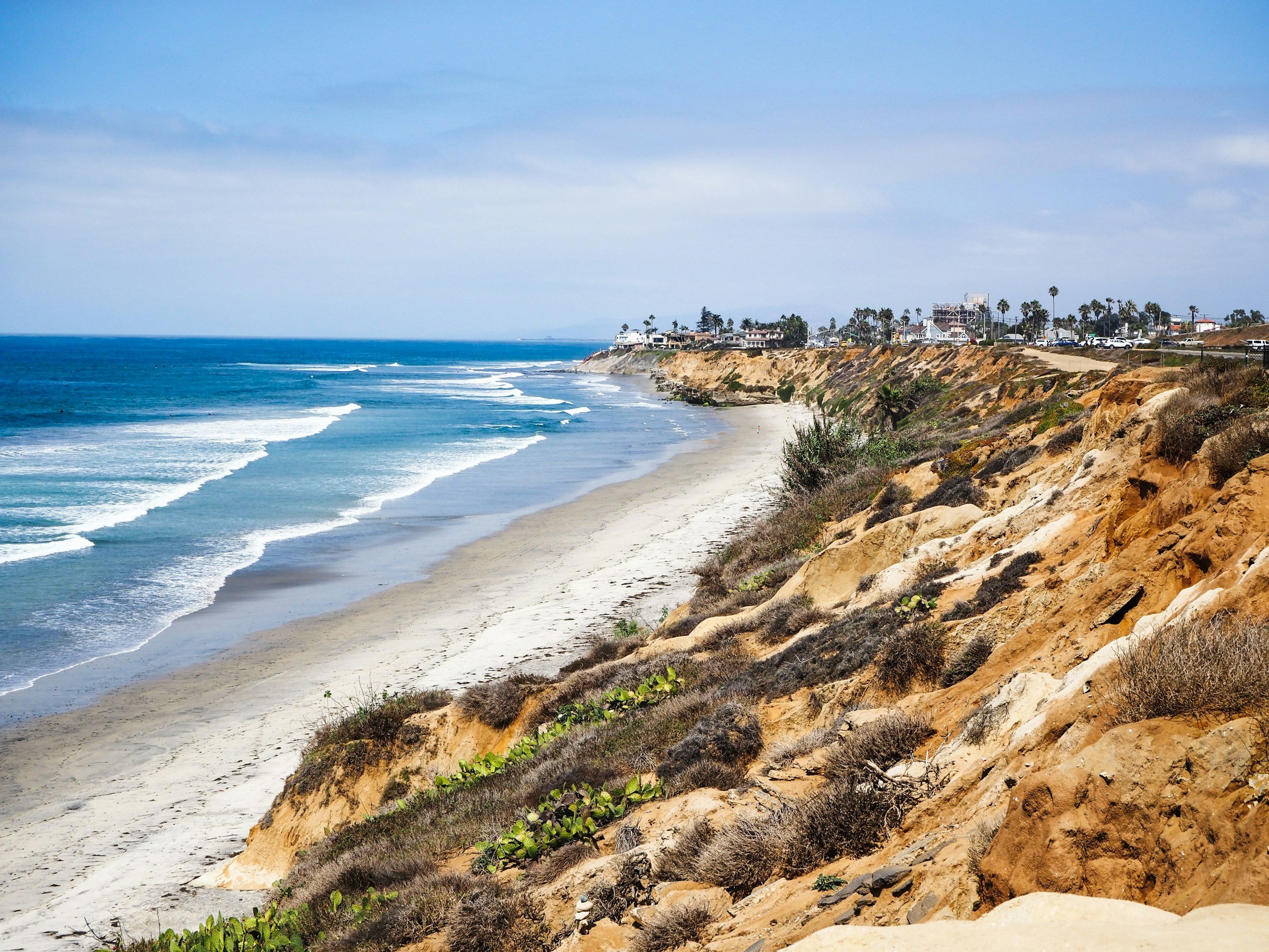 Coastal shoreline with sandy beach, ocean waves, and rocky cliffs with houses and palm trees.