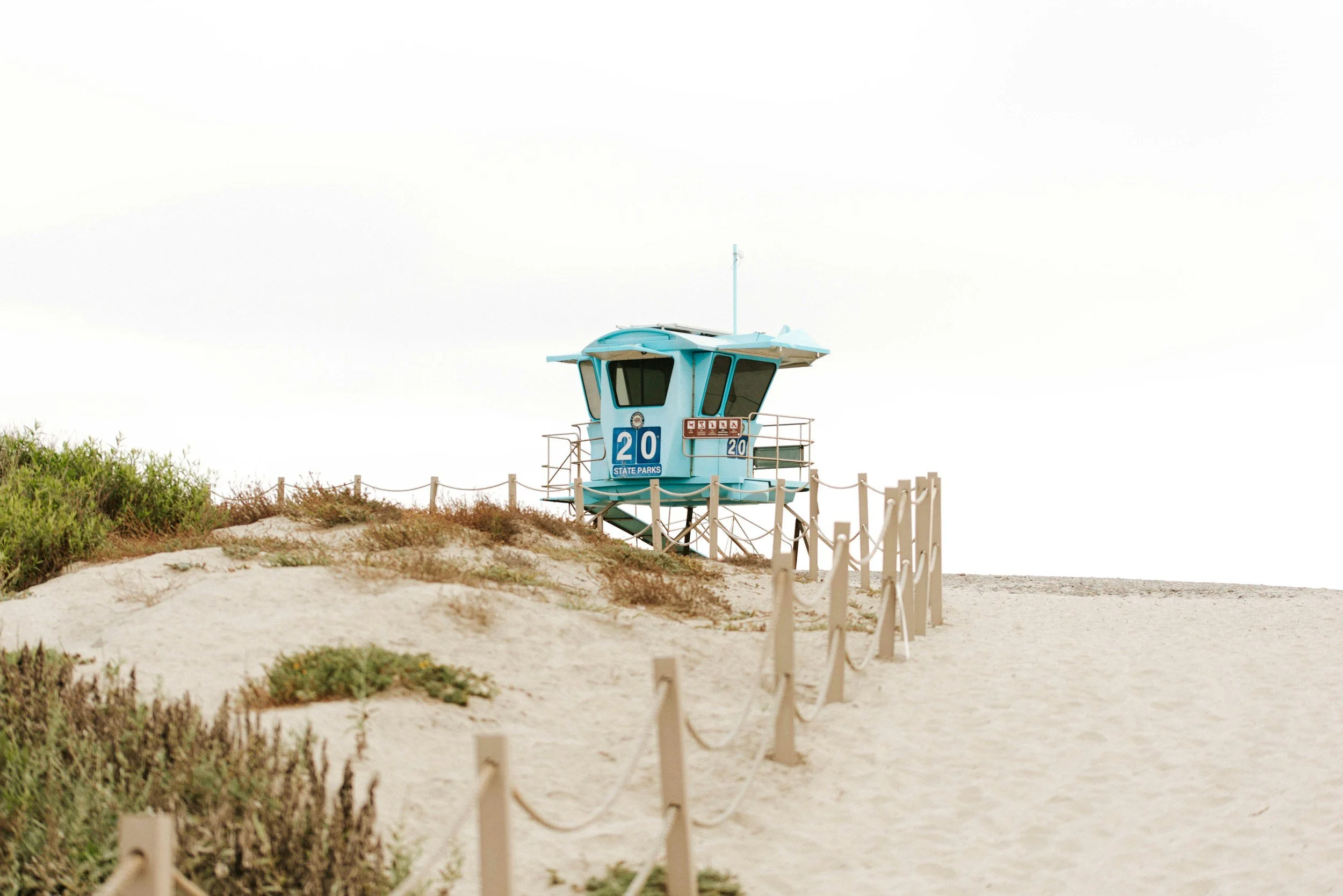 A lifeguard tower on a sandy beach, with a white sky in the background, surrounded by short fence posts and some green bushes.