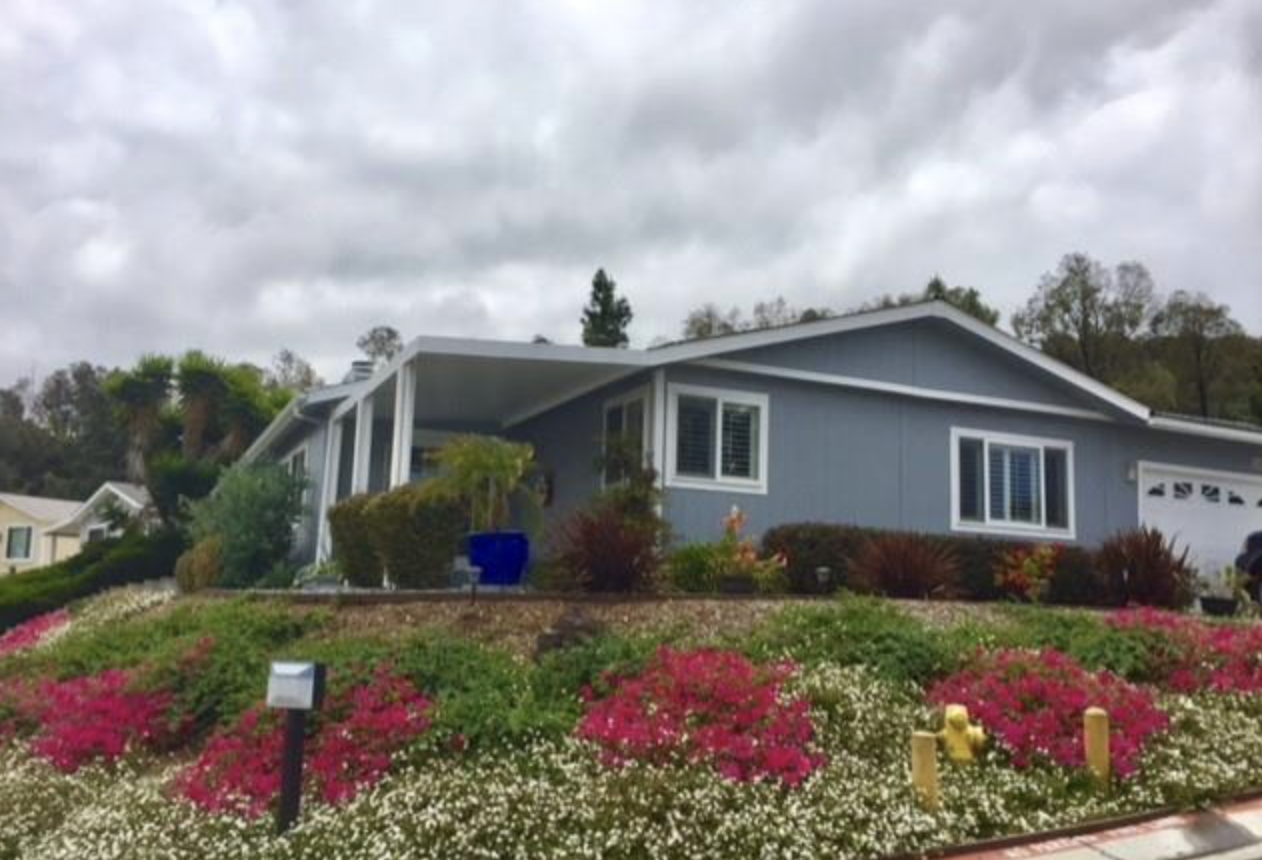 A blue house with a sloped roof, garden with blooming pink and white flowers, bushes, and a cloudy sky overhead.