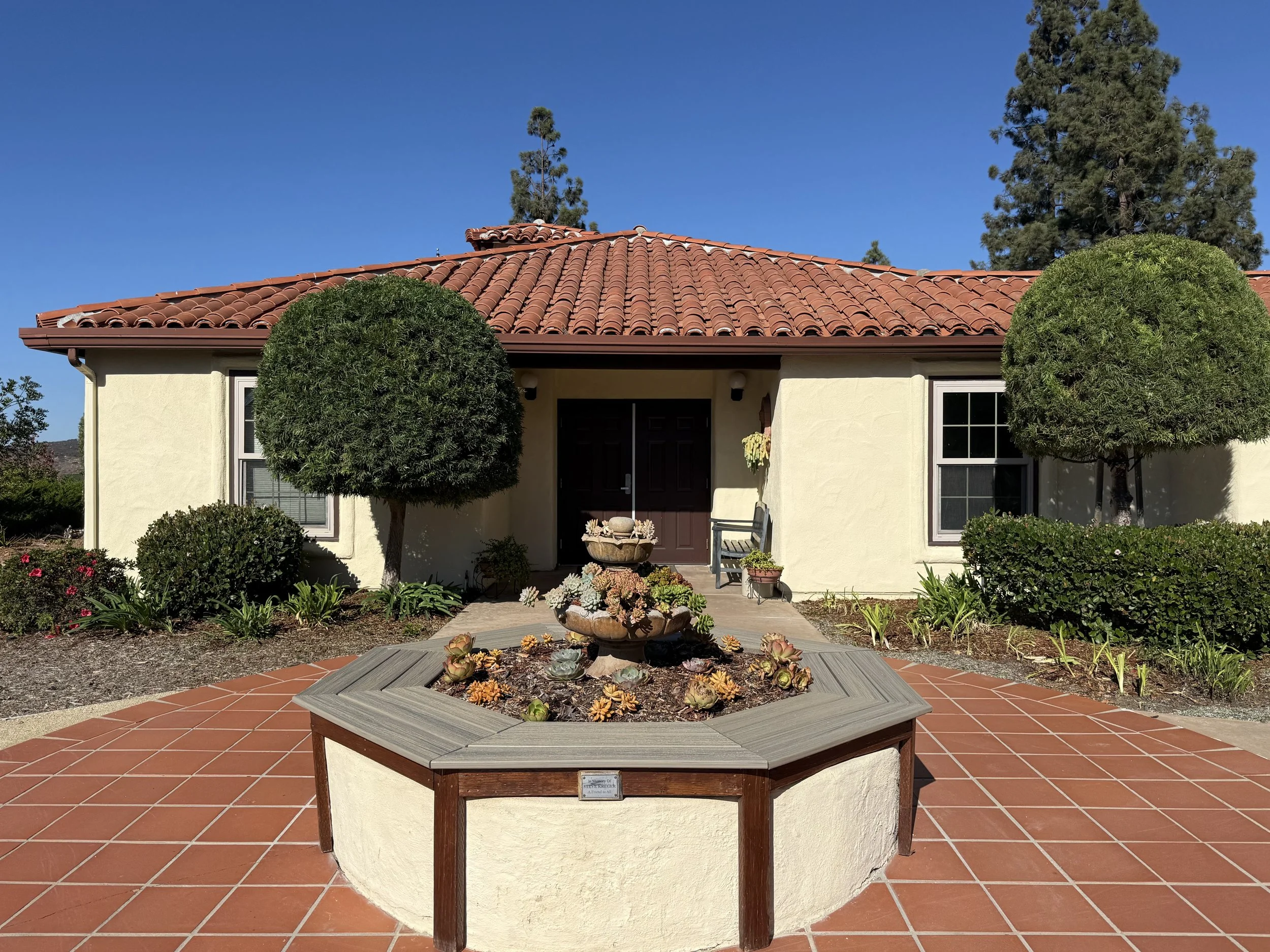 Front yard of a house with a tiled roof, two trees, a patio area with potted plants, and a sculpture in the center surrounded by succulents.