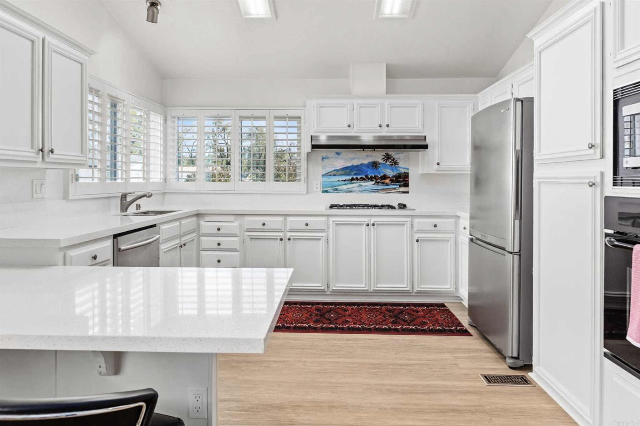 A bright white kitchen with white cabinets, a large window with blinds, a stainless steel refrigerator, and a colorful landscape painting above the stove. There is a red patterned rug on the light wood floor and a white countertop with a sink.