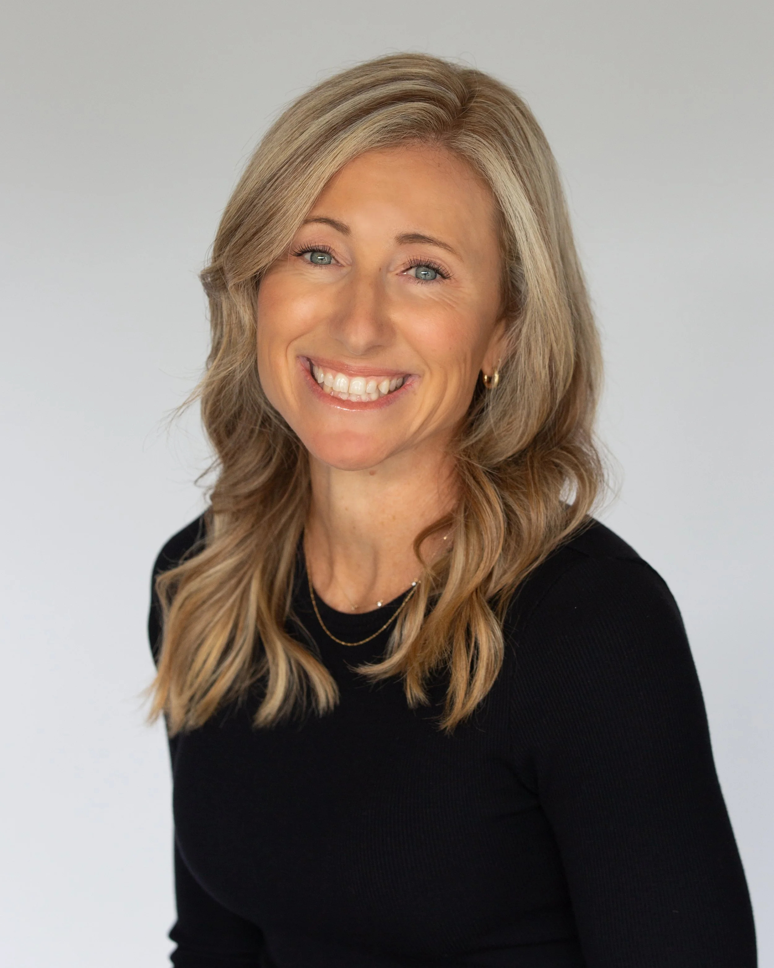 Carlsbad Broker. Close-up of a smiling woman with long, wavy blonde hair, wearing a black top and gold jewelry, standing against a plain light background.