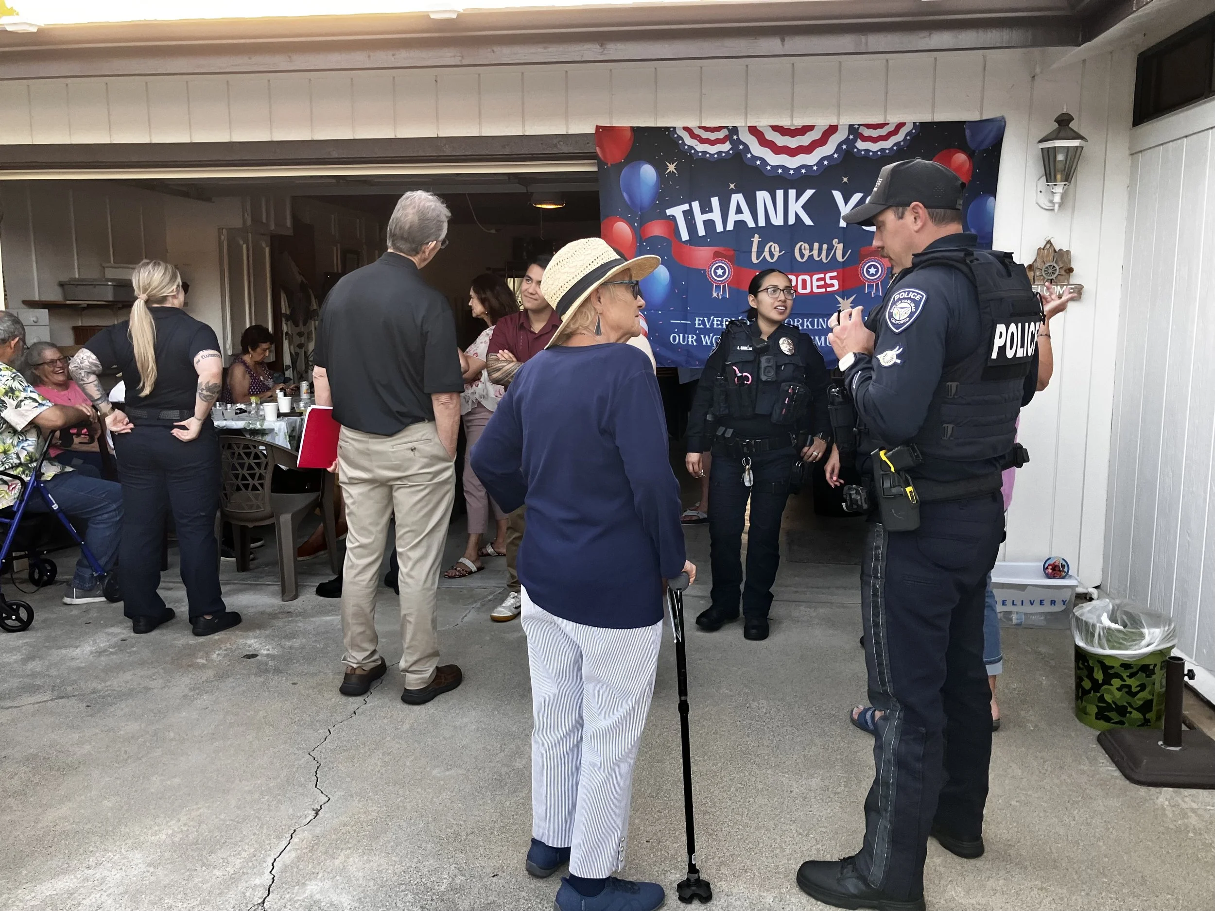 A group of people, including a police officer, gather and chat during a community event with a patriotic theme, featuring a large banner that says 'Thank You to Our Heroes' with red, white, and blue decorations.