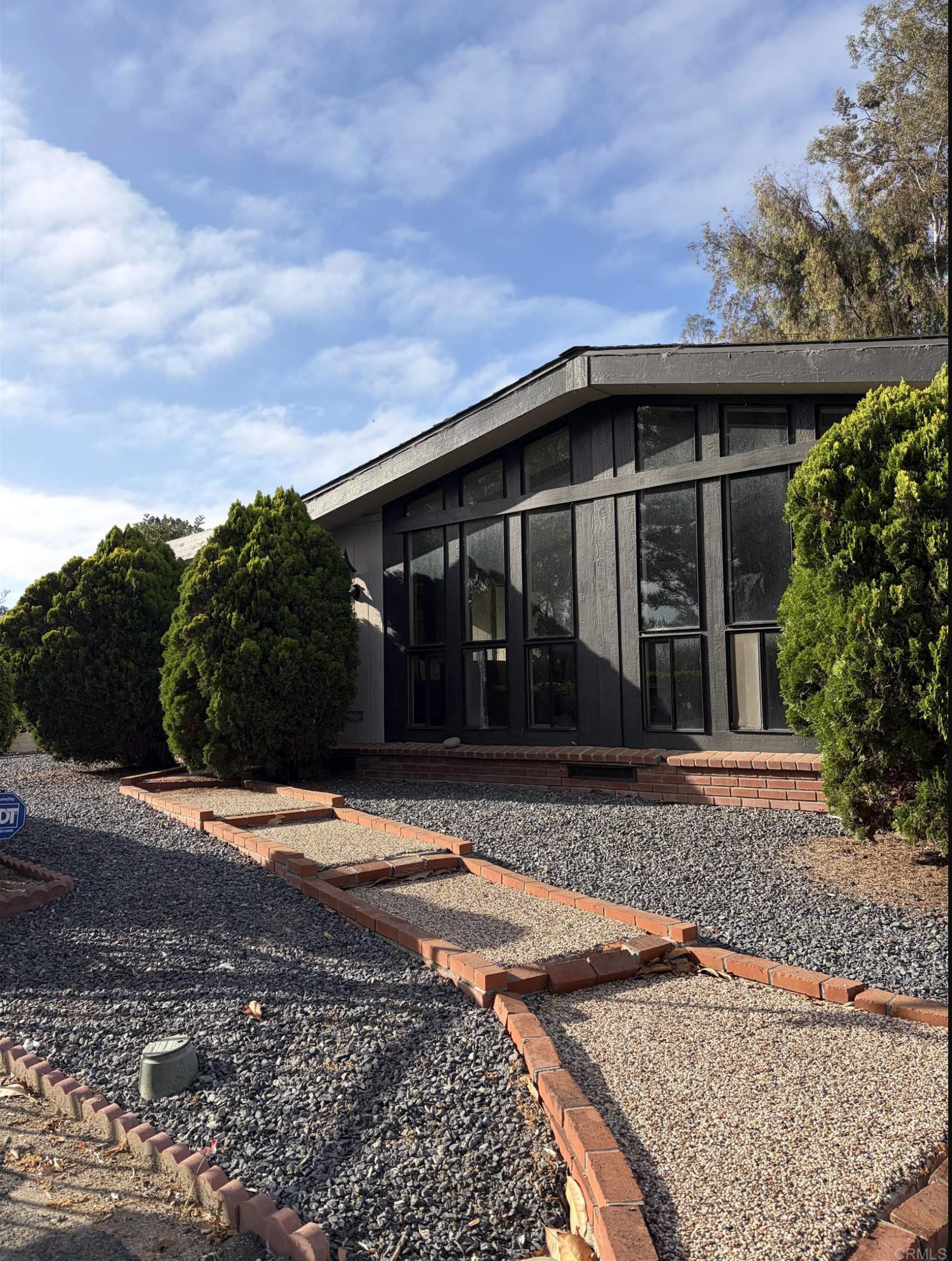 The front yard of a house under construction or renovation, with red brick borders outlining garden beds, a gravel ground cover, green bushes, and a modern house with large windows and a sloped roof. The sky is partly cloudy with some trees visible in the background.