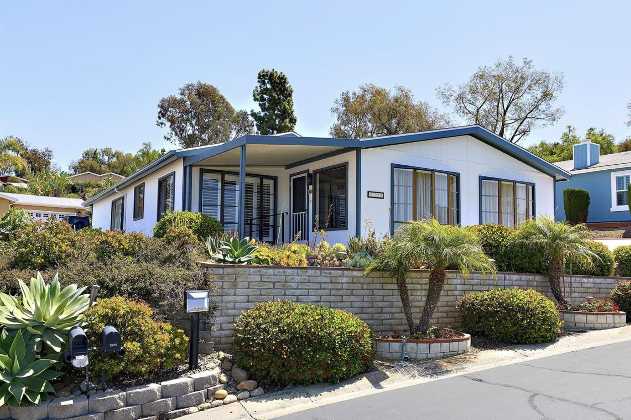 Front yard of a house with a garden featuring succulents and bushes, a brick retaining wall, and mailboxes, with neighboring houses and trees in the background.