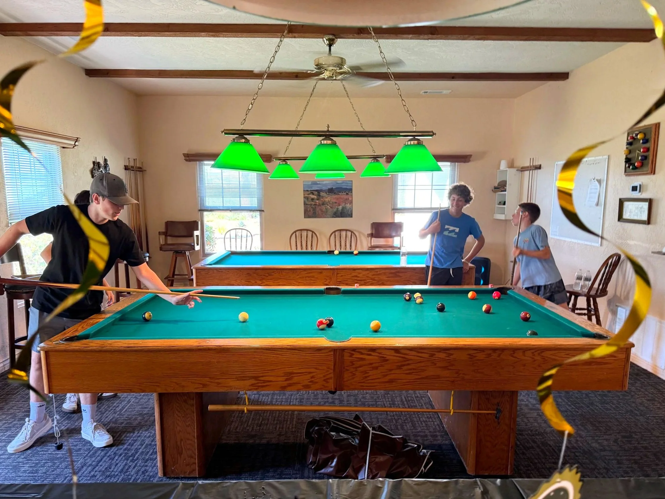 A group of children playing billiards in a cozy room, two of the children are standing by the pool table, one on the left wearing a black shirt and gray cap, and the other on the right wearing a light blue shirt. The room has wooden ceiling beams, large windows with blinds, beige walls, and various decorations, including a framed picture and a whiteboard. The photograph is taken from behind yellow and black streamers hanging from the ceiling.