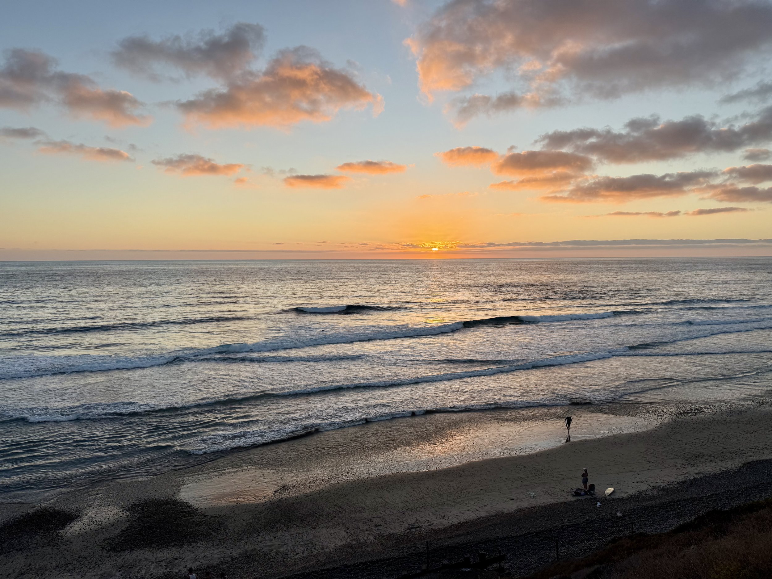 Sunset over the ocean with pink and orange clouds and a person walking along the beach.