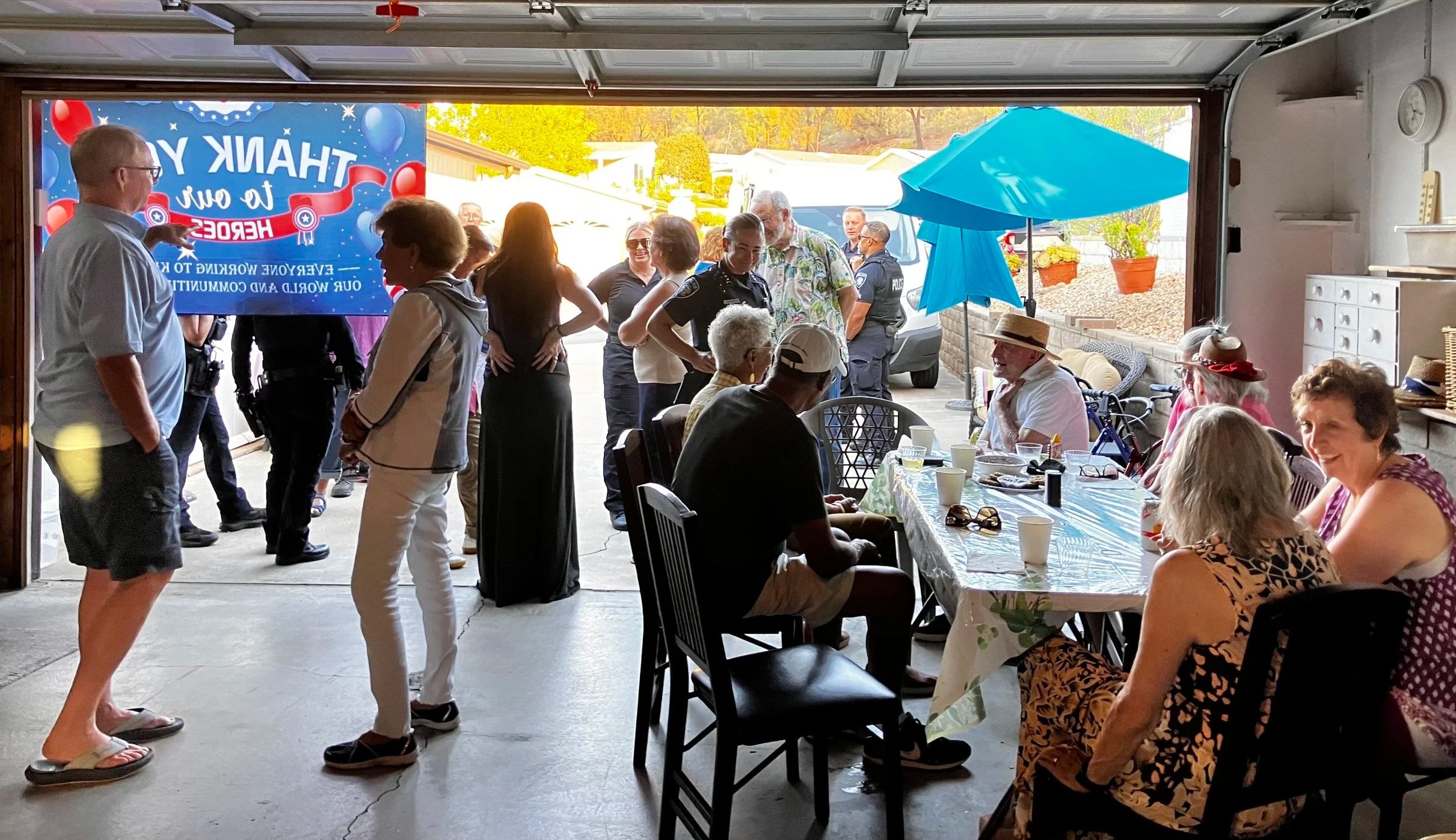 People gathered for a celebration inside and outside a garage, with a large screen displaying 'Thank You to Our Heroes' and a festive setting including tables, chairs, and a blue umbrella.
